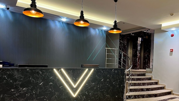 A reception desk with a marble finish and a neon 'V' sign, flanked by two pendant lights. - Ardos Park Hotel (Ankara)