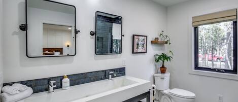 Un baño con un lavamanos grande blanco, un espejo, un inodoro y una ventana con vistas a árboles.