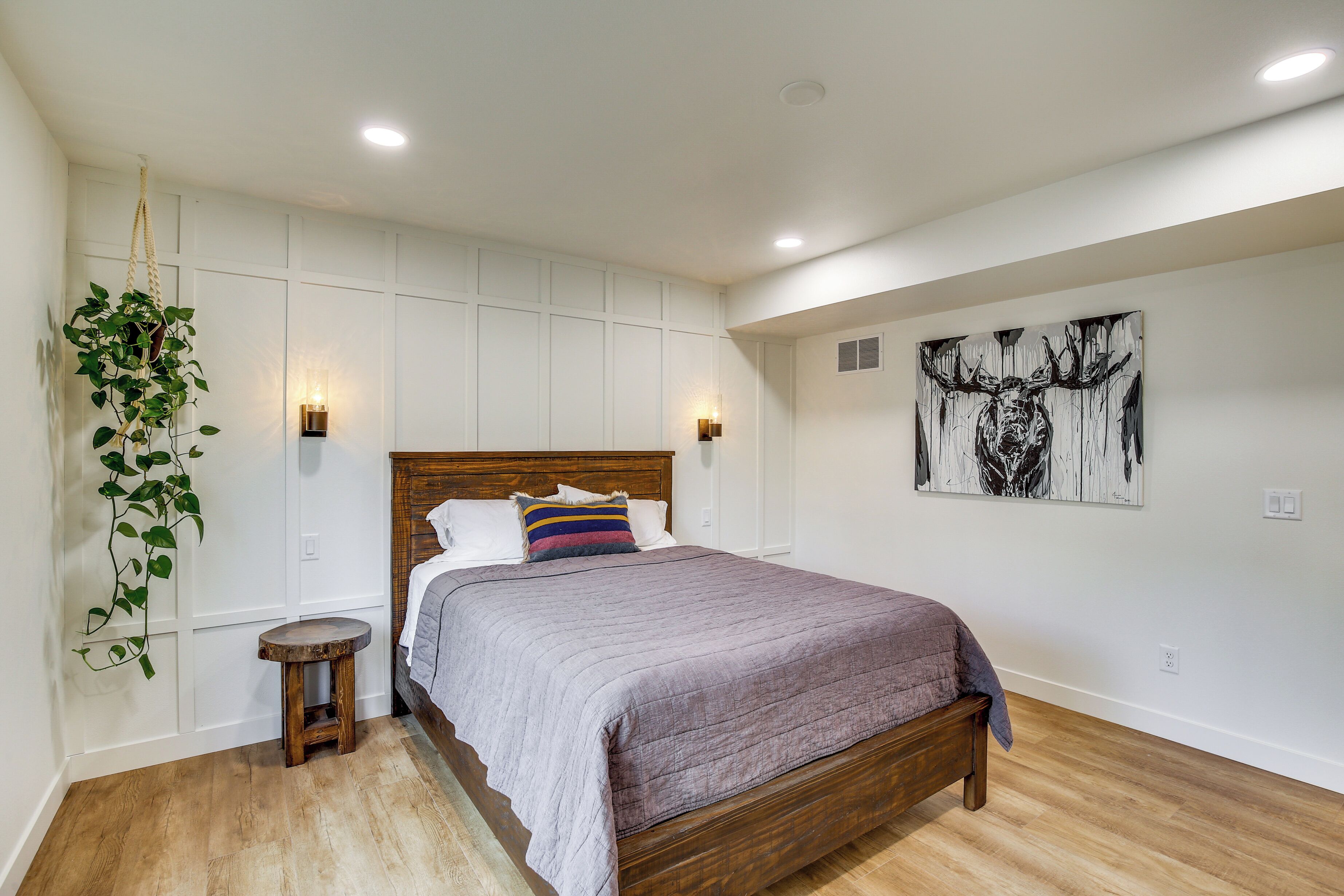 A bedroom with a wooden bed, a hanging plant, a small stool, and a wall-mounted light fixture.