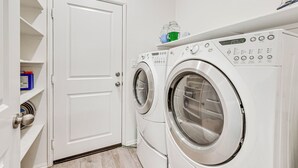 Uma lavanderia com duas máquinas de lavar de abertura frontal, uma prateleira com produtos de limpeza e uma porta.