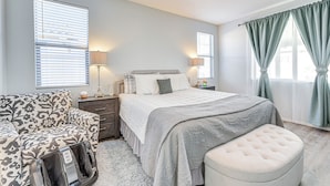 Quarto com cama, mesas de cabeceira, abajur, cadeira e janela com cortinas.