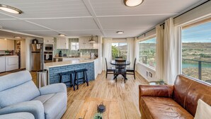 Una cocina moderna con una isla central, un comedor con mesa y sillas, una sala de estar con sofá y ventanas amplias que ofrecen vistas al paisaje.
