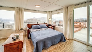 Un dormitorio con una cama grande, un escritorio, una silla y vistas a las montañas a través de las ventanas.