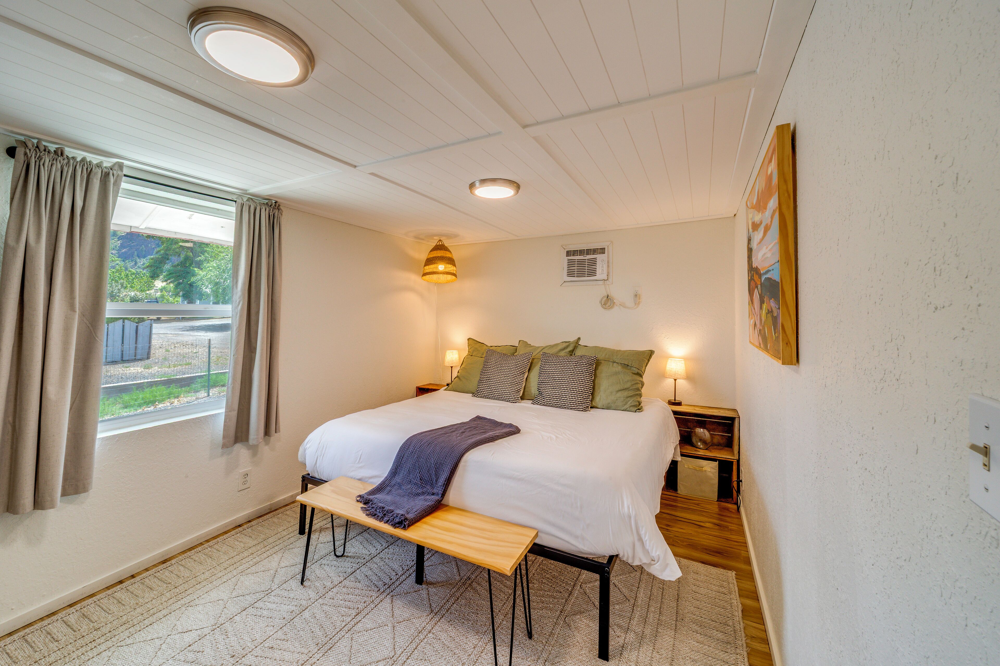 Une chambre à coucher avec un lit, des lampes de chevet, un banc, une fenêtre avec des rideaux et un tableau au mur.