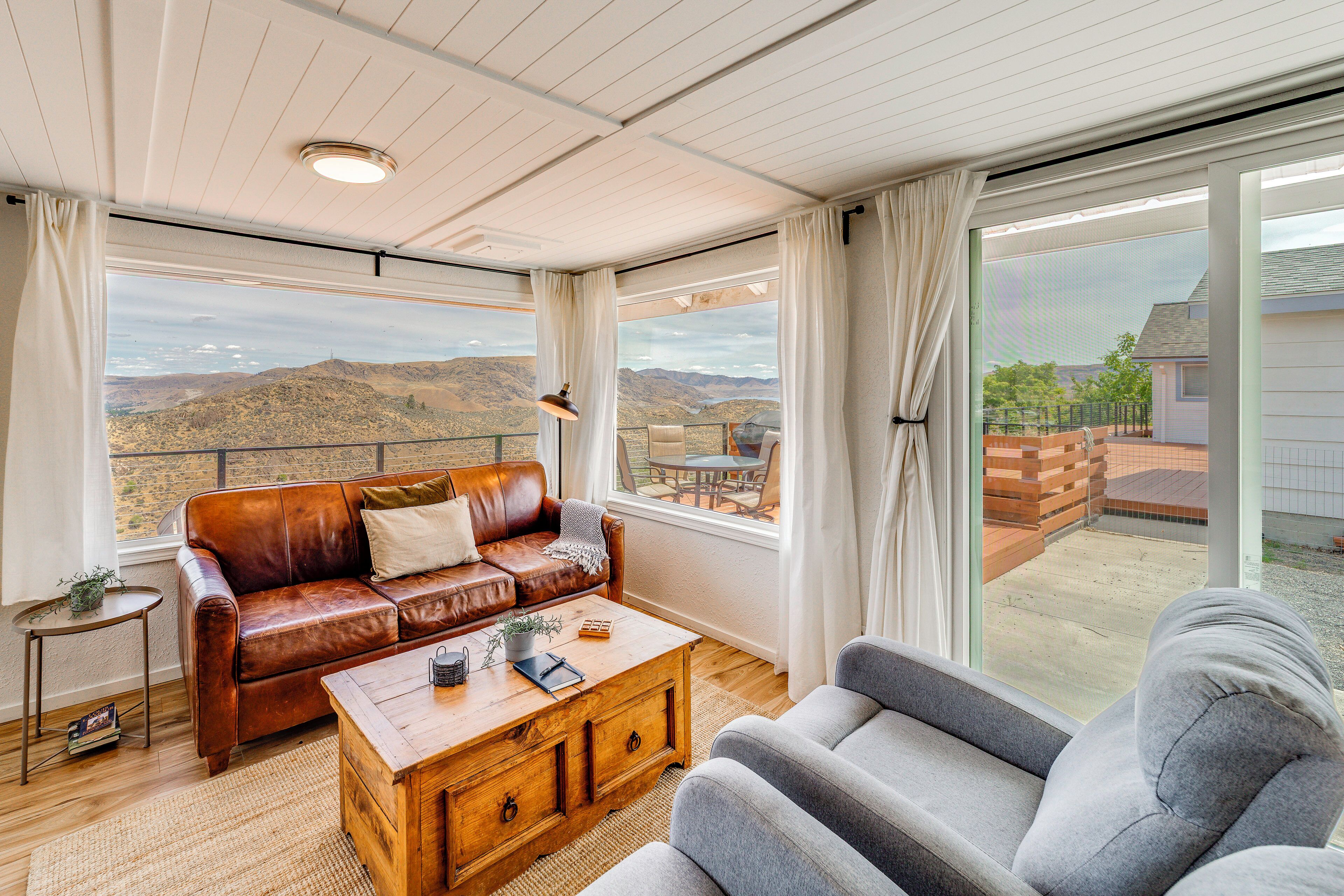 A spacious living area with a leather sofa, a wooden coffee table, and a gray armchair. Large windows offer a view of the mountains.
