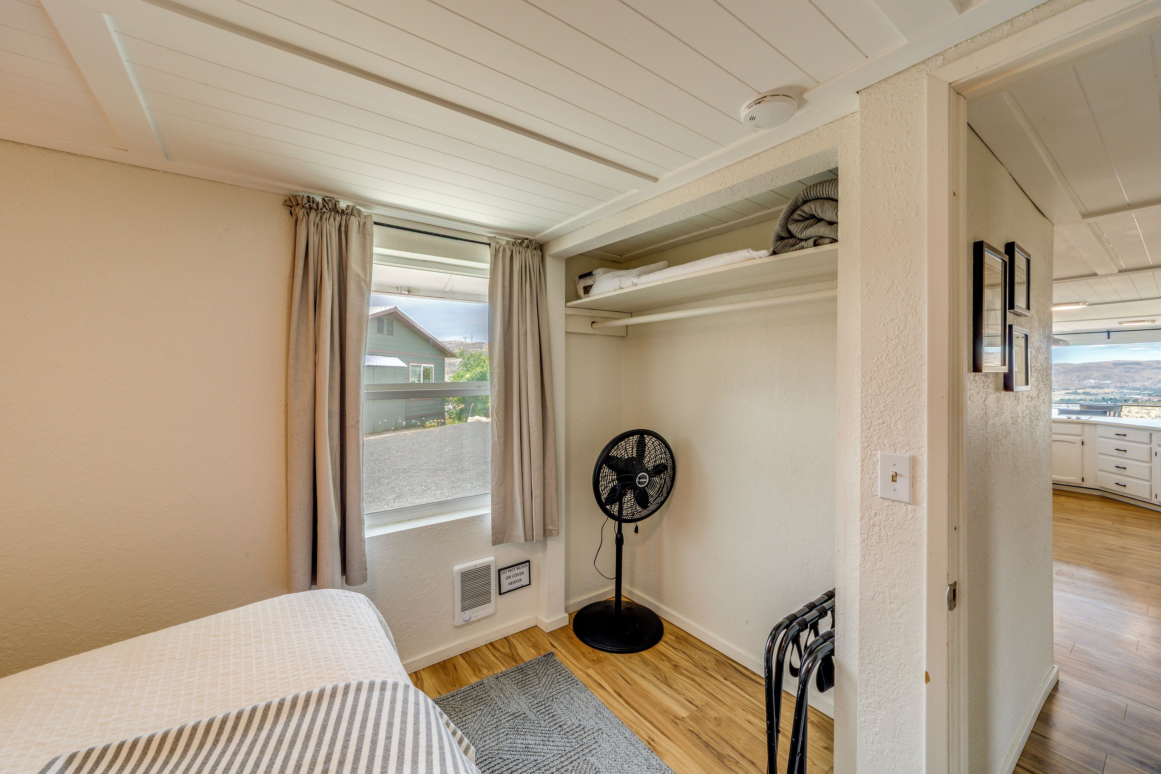 A bedroom with a bed, a window with curtains, a standing fan, and a view of the outside.