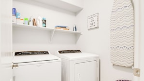 Une buanderie équipée de deux machines à laver et deux sèche-linge blancs, d’étagères contenant des produits de nettoyage et d’un panneau affiché au mur.