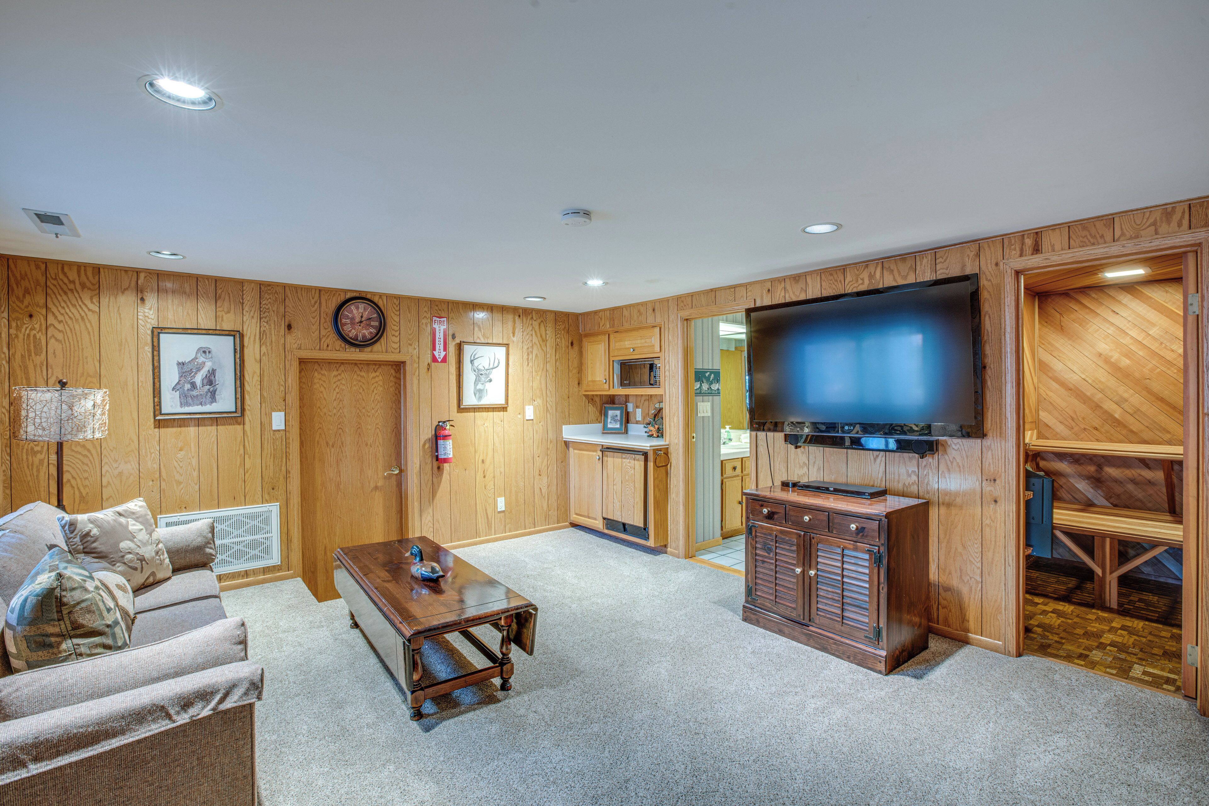 A wooden-paneled living room with a television, a sofa, a coffee table, and a kitchen area in the background.