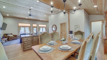 A dining area with a wooden table set for four, surrounded by chairs. A chandelier hangs above the table, and there are windows with curtains on the wall.