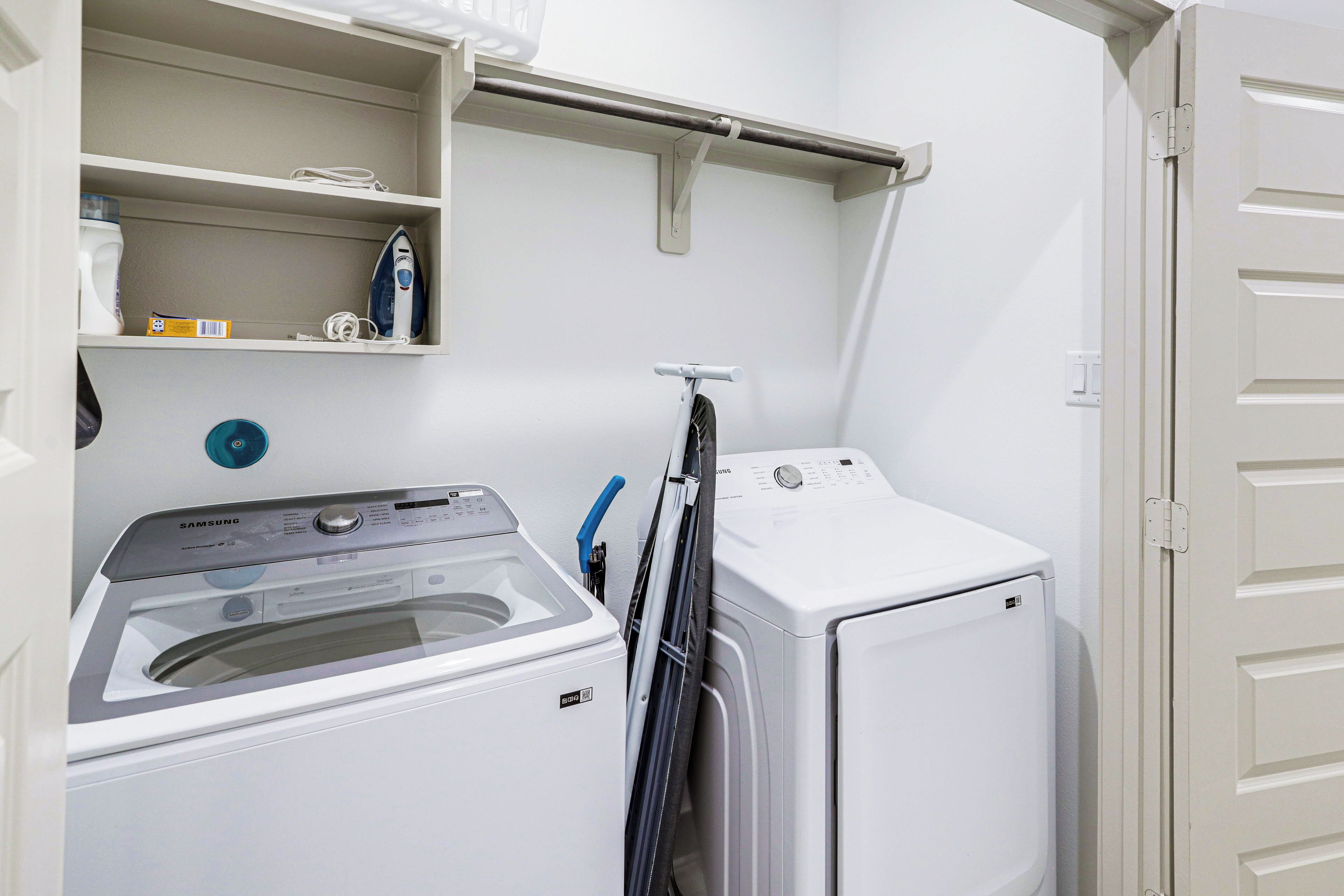 Une buanderie équipée d’un lave-linge et d’un sèche-linge à chargement frontal, d’une étagère contenant des produits de lessive et d’une porte située sur le côté droit.