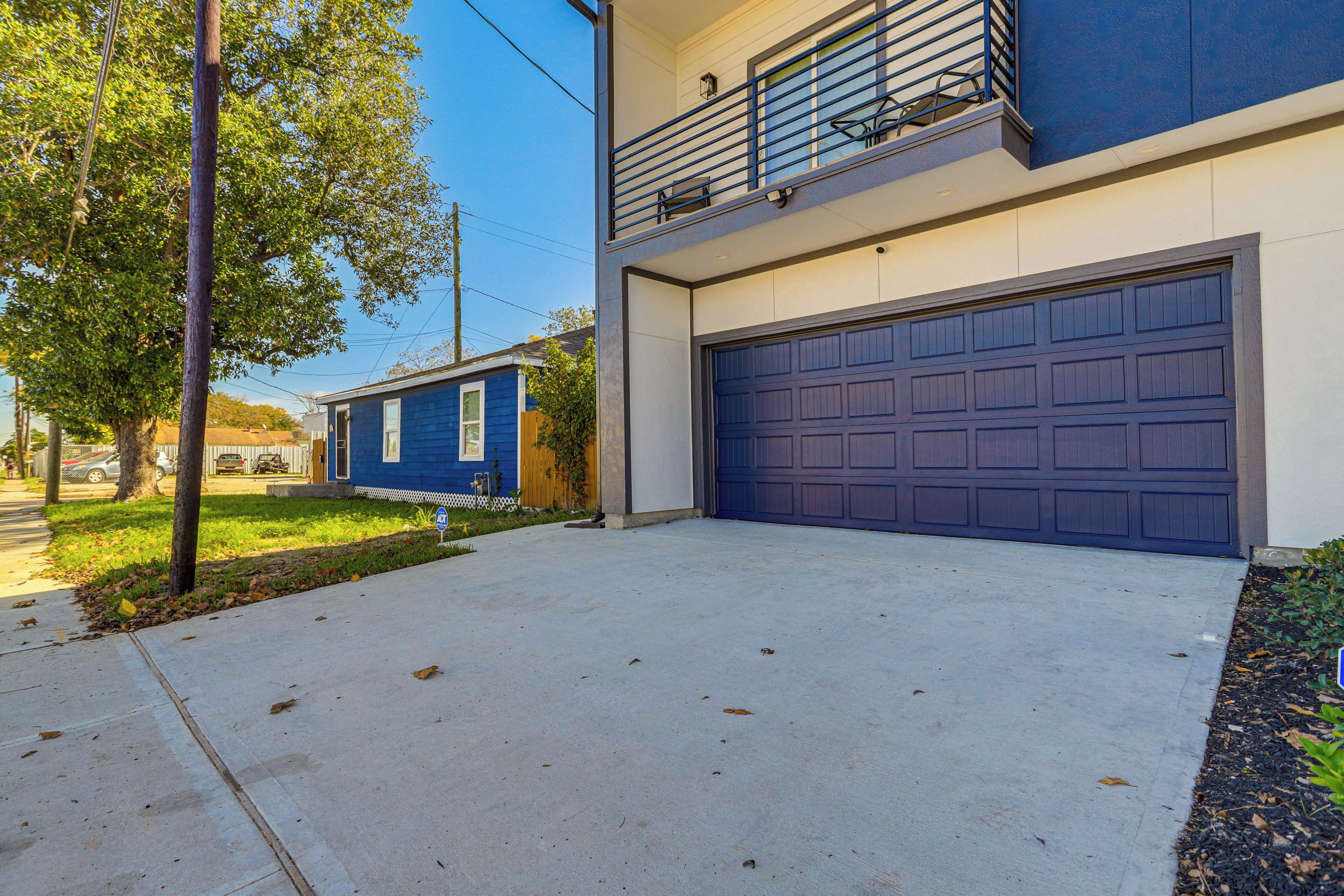 A two-story house with a blue garage door, a balcony with a metal railing, and a tree in the front yard.