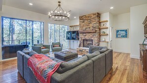 A spacious living room with a large sectional sofa, a stone fireplace, and a large window overlooking a forest. - Waterfront Home w/ 3 Decks, Fire Pits and Hot Tub! (Terry)