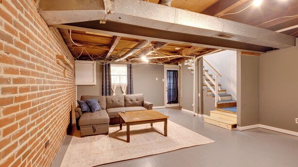 A spacious basement with exposed brick walls, a wooden ceiling, a sofa, a coffee table, and a staircase leading upstairs. - Charming State College Home - 4 Mi to Psu! (State College)