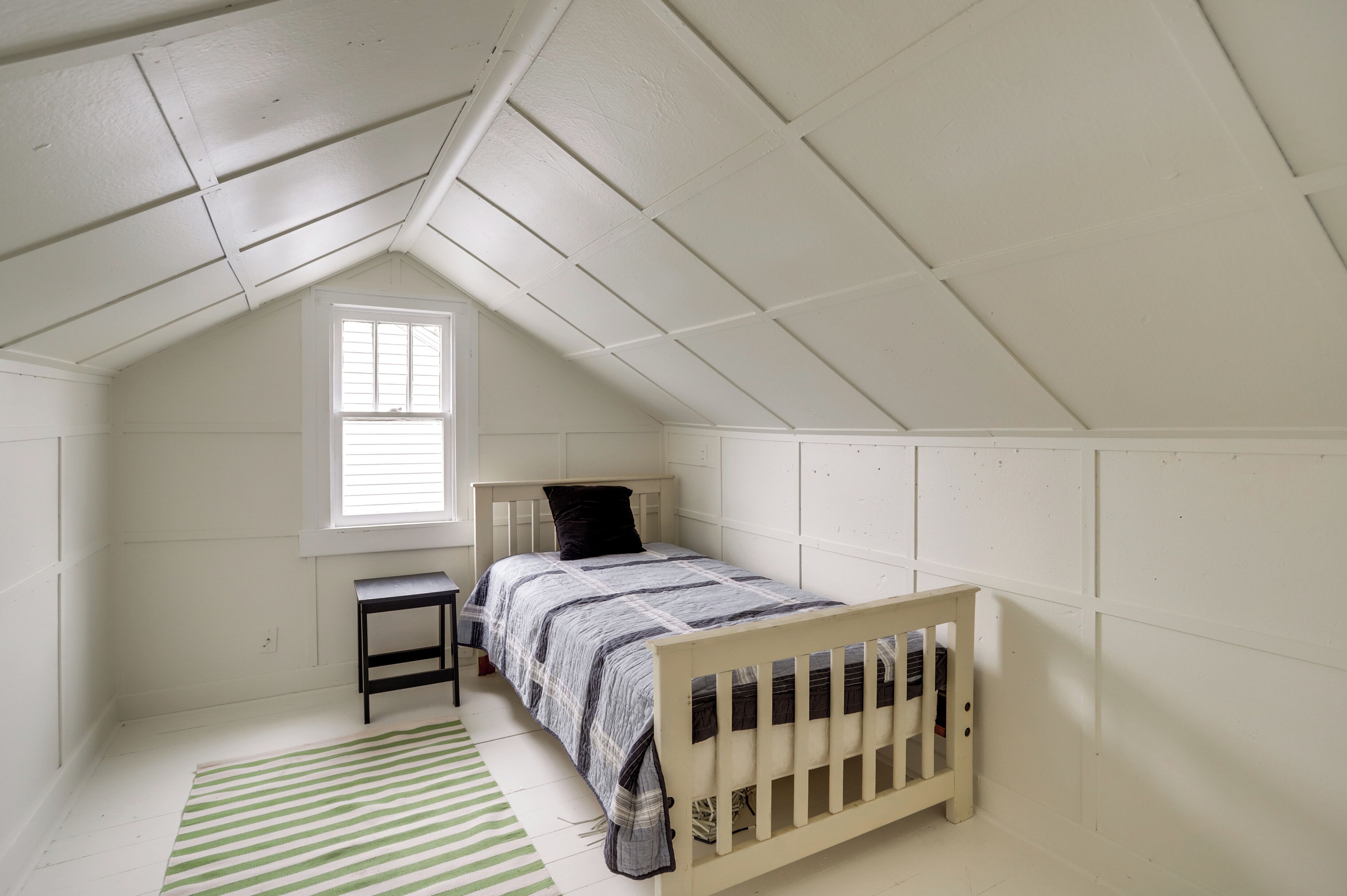 Une petite chambre mansardée, aux murs blancs, avec un lit simple, une fenêtre et un tapis à rayures vertes.