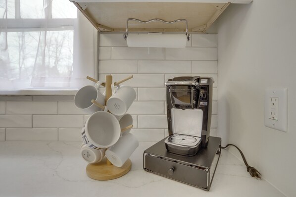 Uma cozinha com uma cafeteira, uma pilha de chávenas brancas e uma janela com vista para árvores.