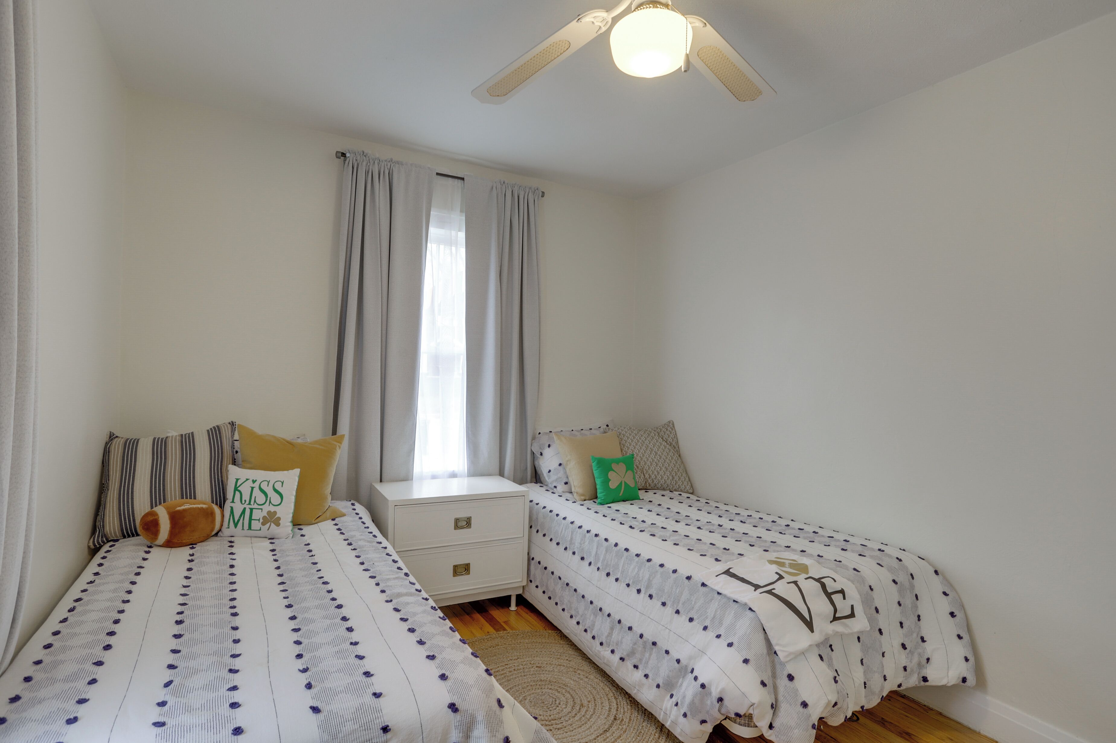 A bedroom with two single beds, a nightstand, a window with curtains, and a ceiling fan.