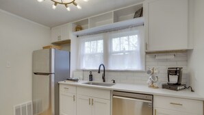 Una cocina moderna con armarios blancos, un refrigerador de acero inoxidable, lavavajillas empotrado, una ventana con cortinas translúcidas y una cafetera sobre la encimera.
