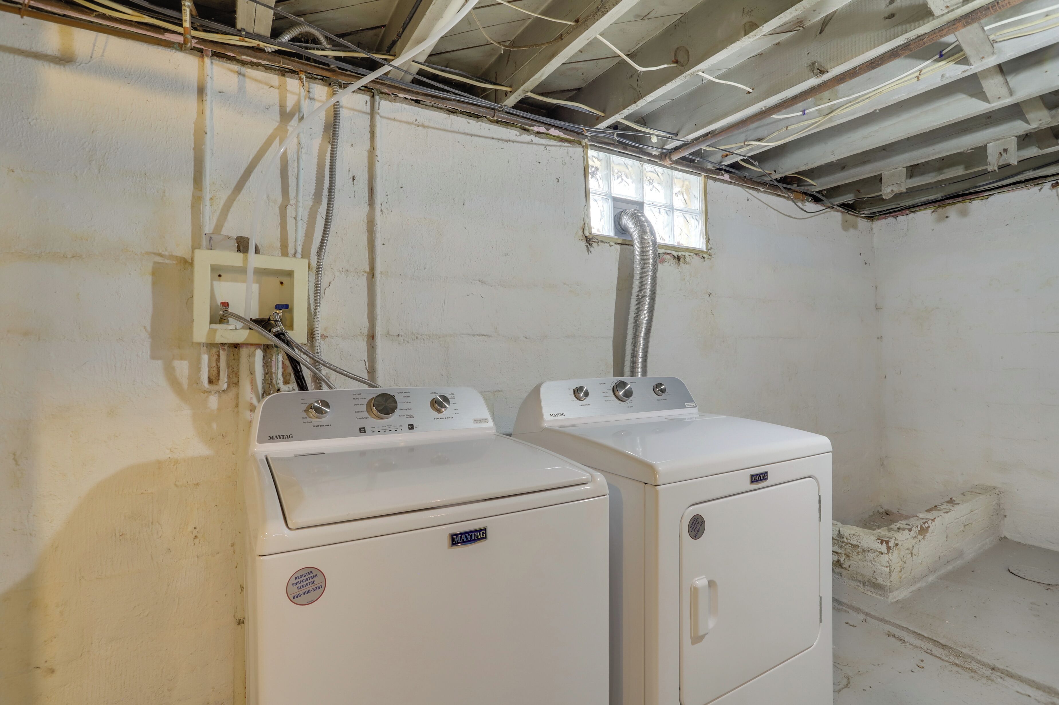 A laundry room with two Maytag washers and dryers, exposed ceiling wiring, and a window with a view of the outside.