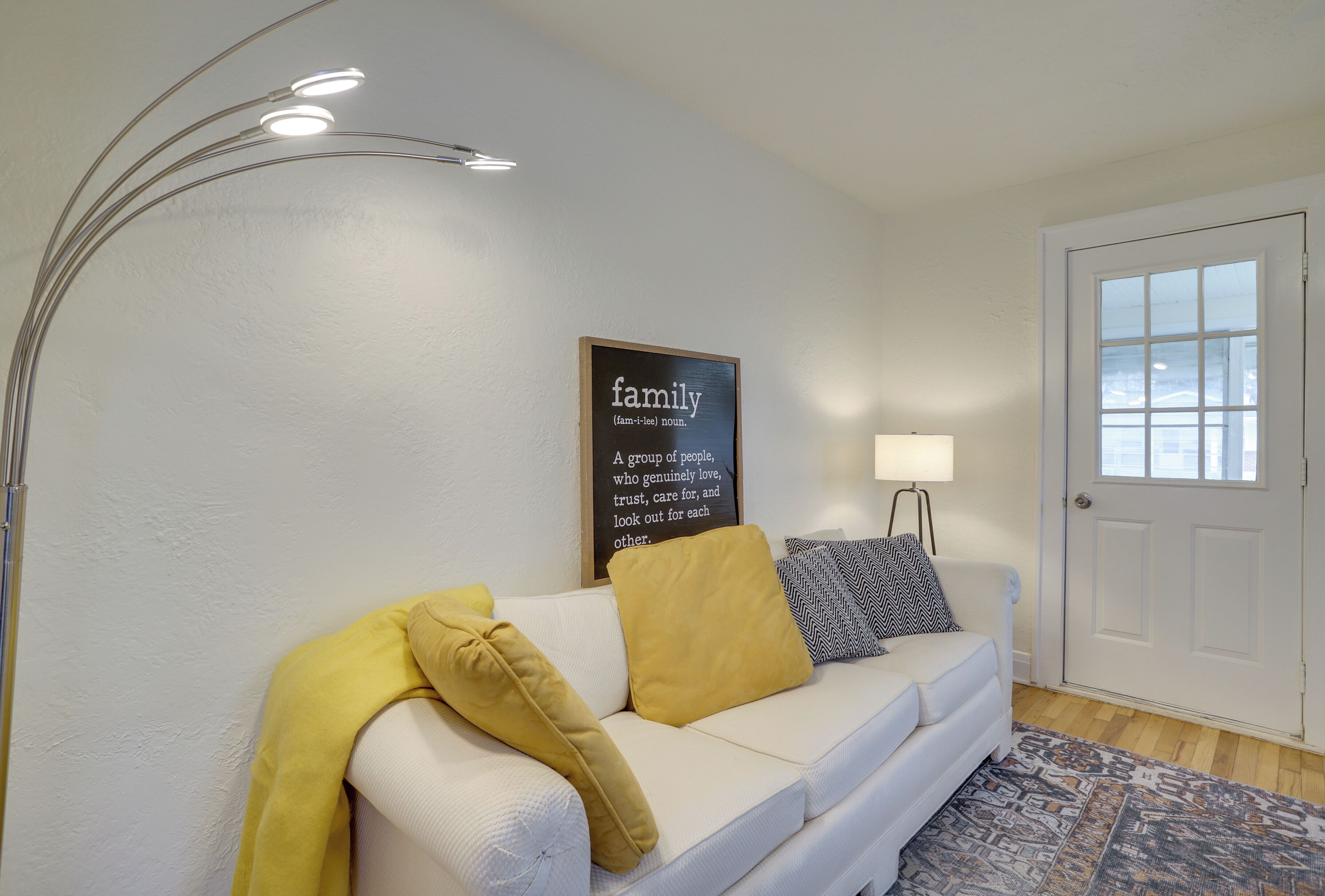 A living room with a white sofa, yellow and black pillows, a patterned rug, a wall-mounted lamp, and a framed sign.