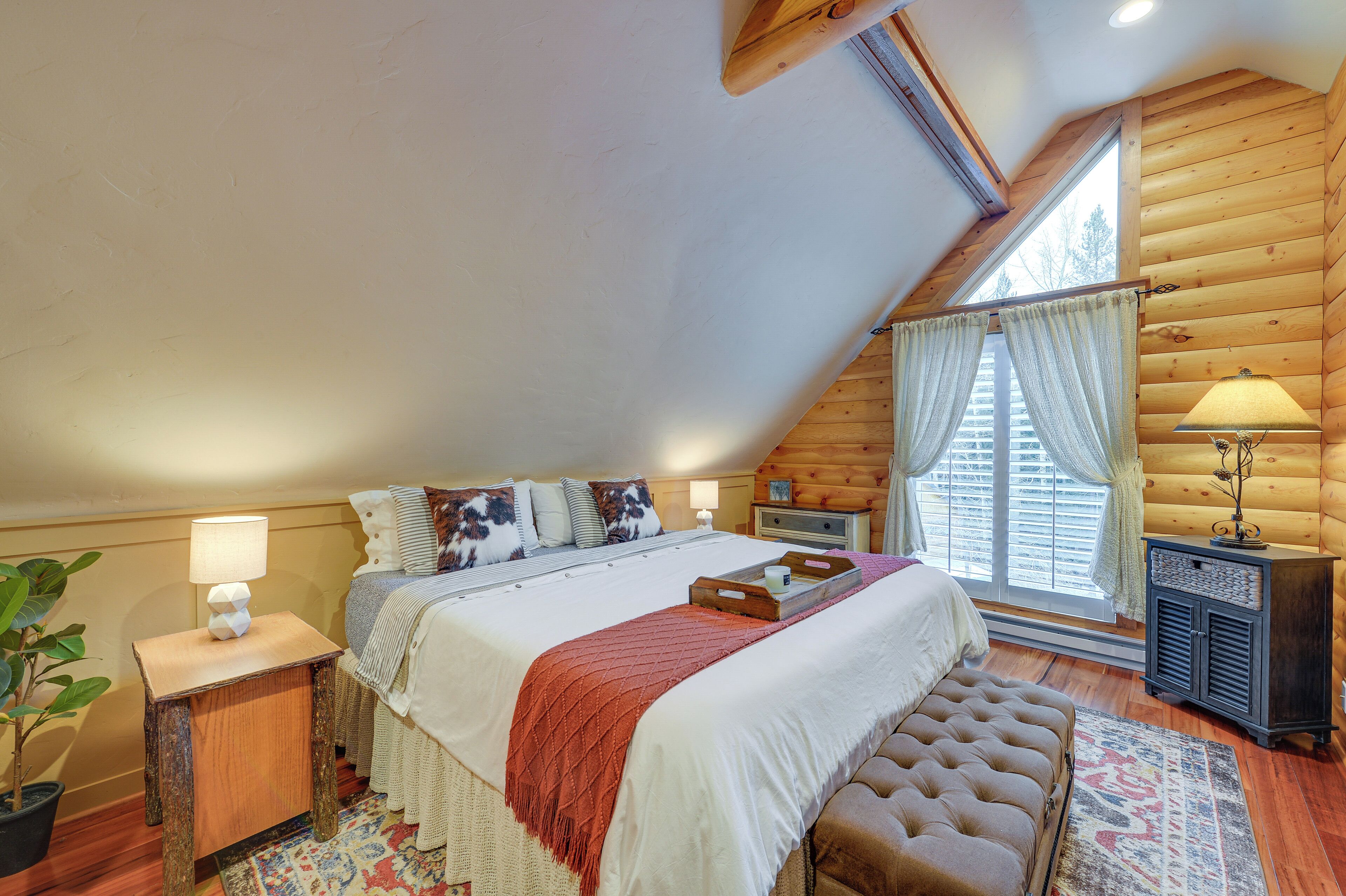 A cozy bedroom with a large bed, a bench, a nightstand with a lamp, and a window with curtains.