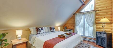 A cozy bedroom with a large bed, a bench, a nightstand with a lamp, and a window with curtains.