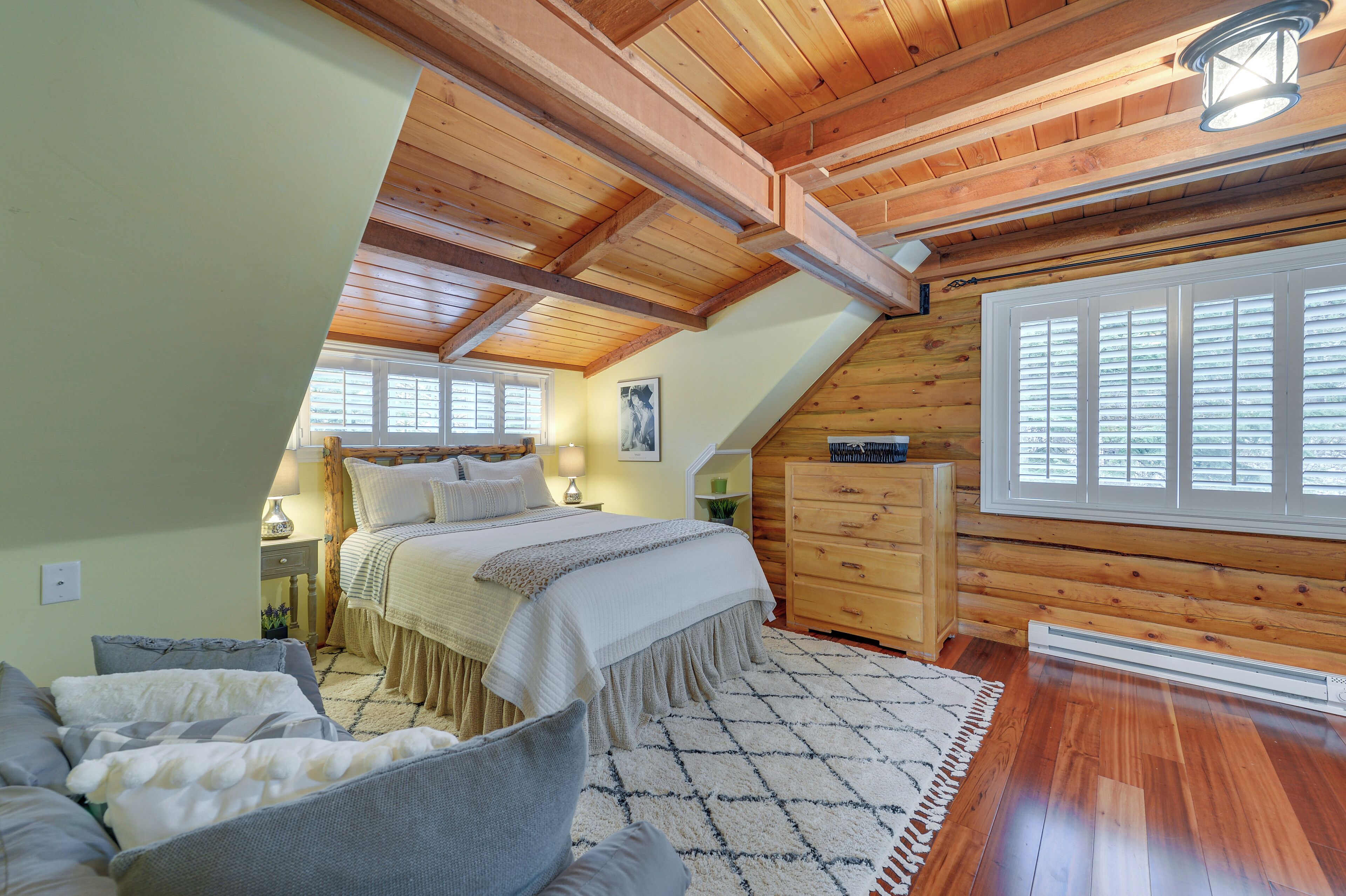 Un dormitorio con techo inclinado, vigas de madera, una cama con cubrecama blanco, una mesita de noche, un aparador y un ventanal grande con persianas.