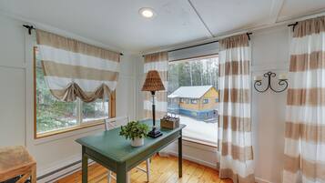 Quarto com piso de madeira, uma mesa verde com uma planta, uma luminária e uma janela com cortinas listradas.