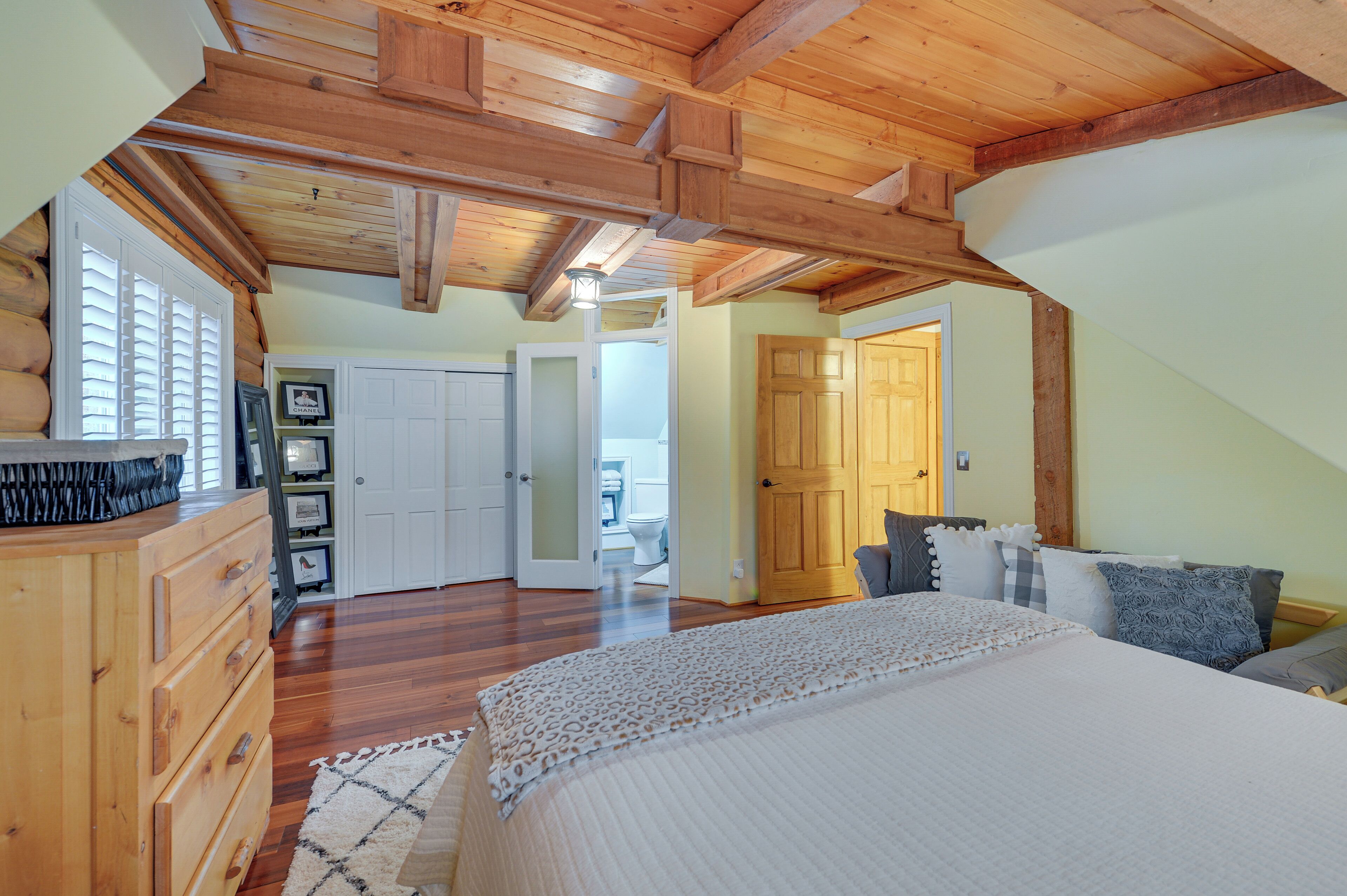 A bedroom with a large bed, wooden ceiling beams, a dresser, and a door leading to a bathroom.