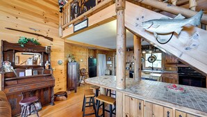 A wooden cabin interior with a piano, bar stools, a fish mounted on the wall, and a kitchen area. - Rustic Fancy Gap Cabin w/ Blue Ridge Parkway Views (Fancy Gap)