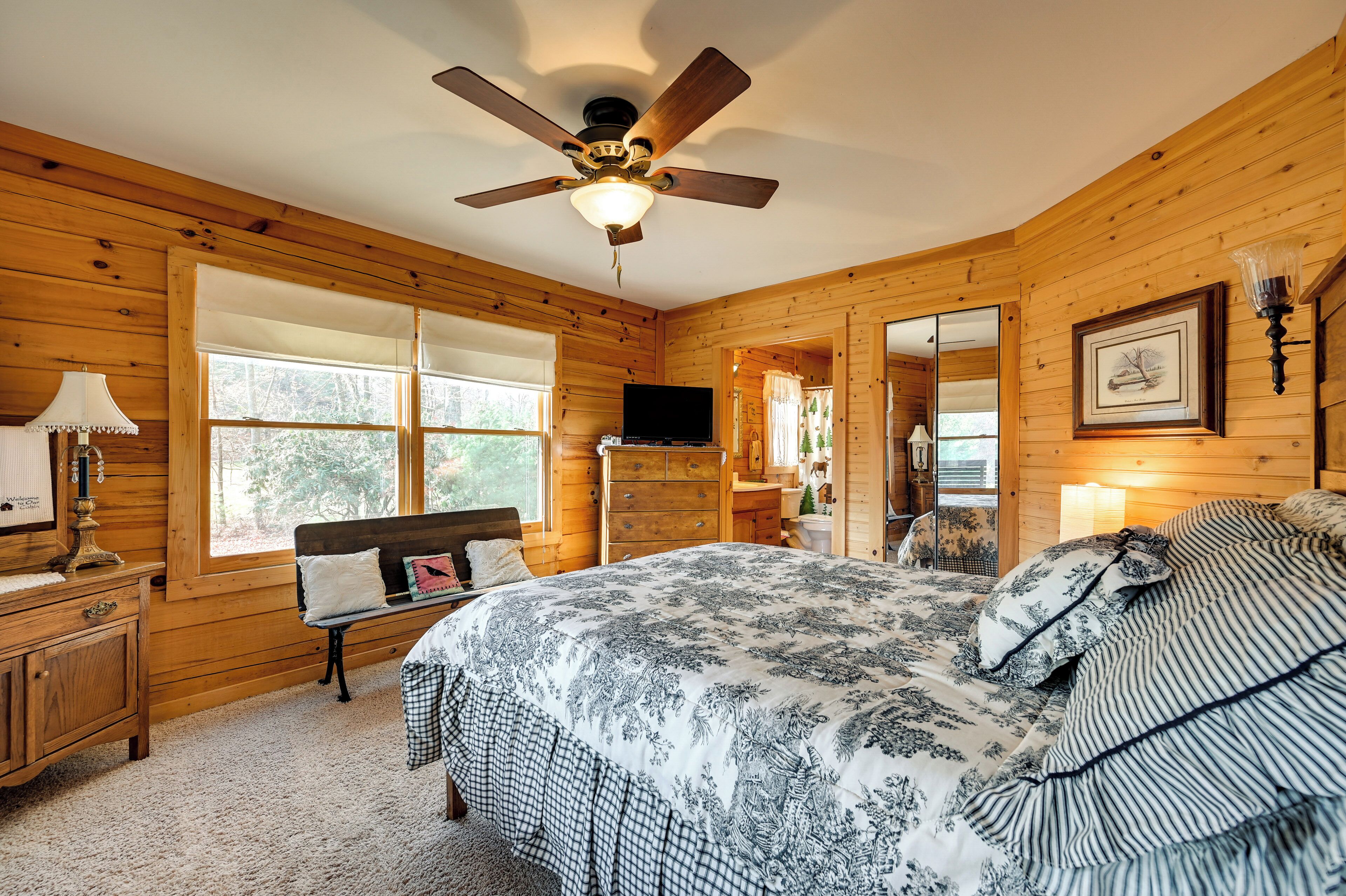 A wooden cabin bedroom with a bed, a bench, a dresser, a television, and a ceiling fan.