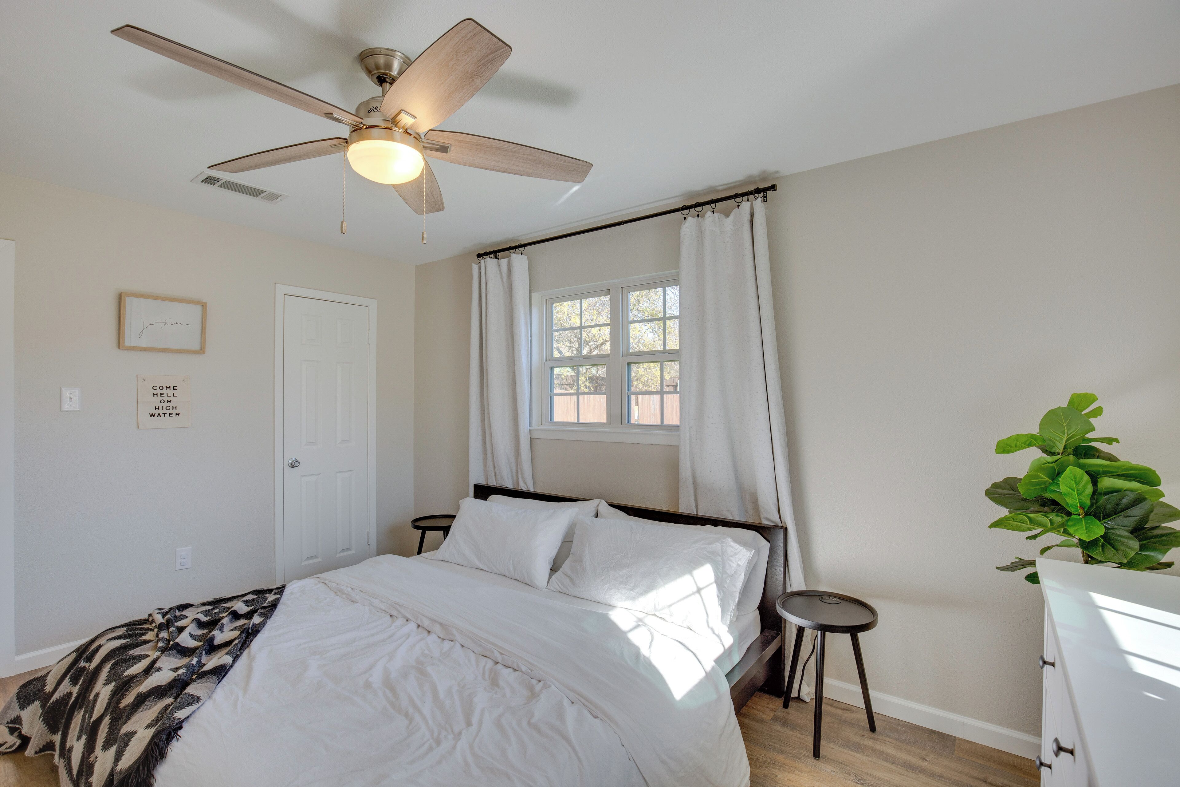 A bedroom with a bed, a ceiling fan, a window with curtains, a bedside table, and a potted plant.