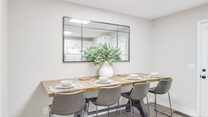 A dining area with a long wooden table, four grey chairs, a white vase with green foliage, and a large mirror above the table. - Modern Townhome: Walk to Tennessee Tech University (Cookeville)