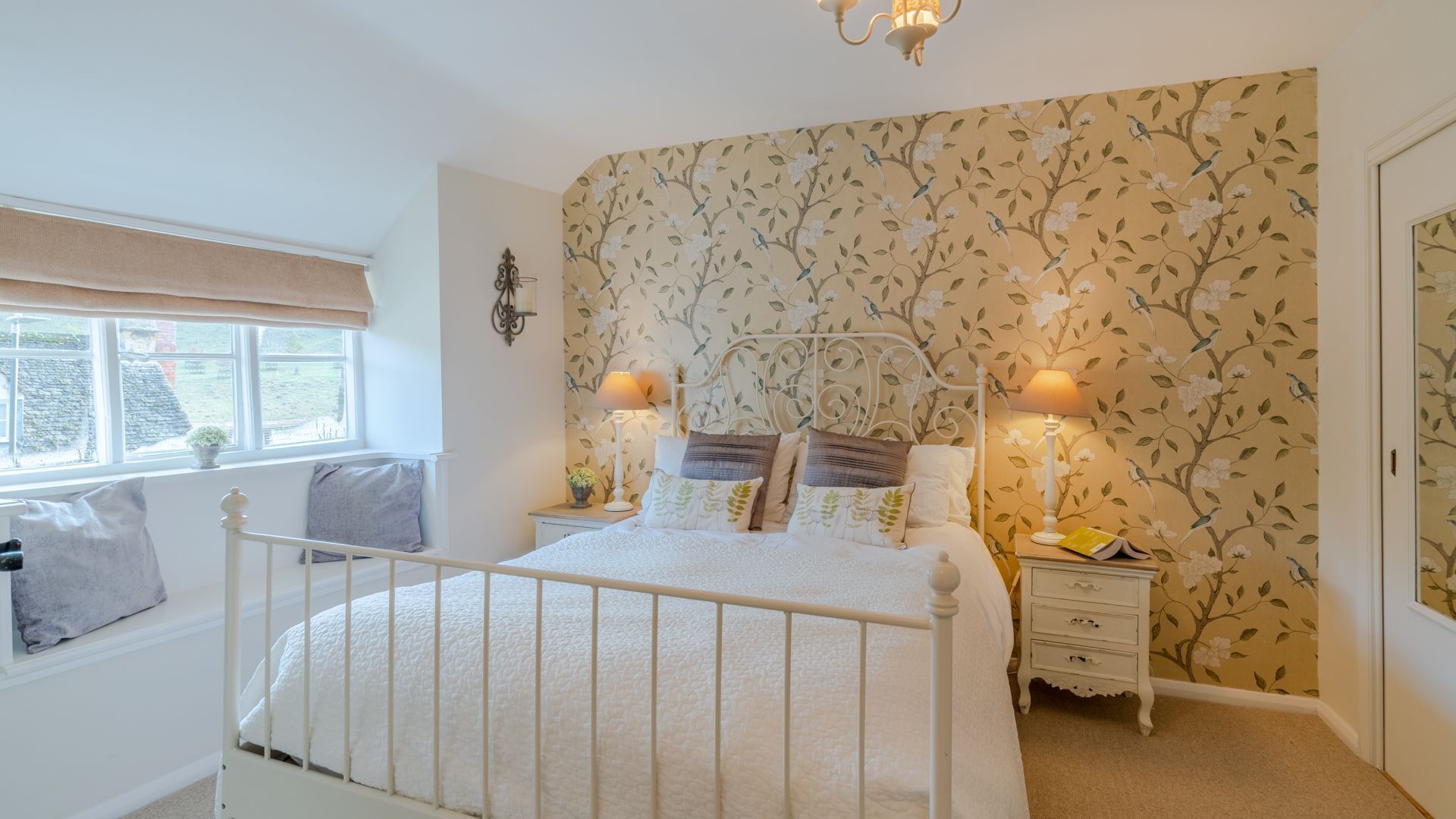 A bedroom with a white bed, two bedside tables with lamps, a window with a view, and floral wallpaper.