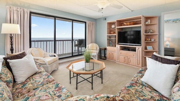 A living room with a large window, a flat-screen TV, wicker furniture, and a glass coffee table.