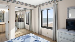A hotel room with a bed, a TV, a dresser, and a window with a beach view.