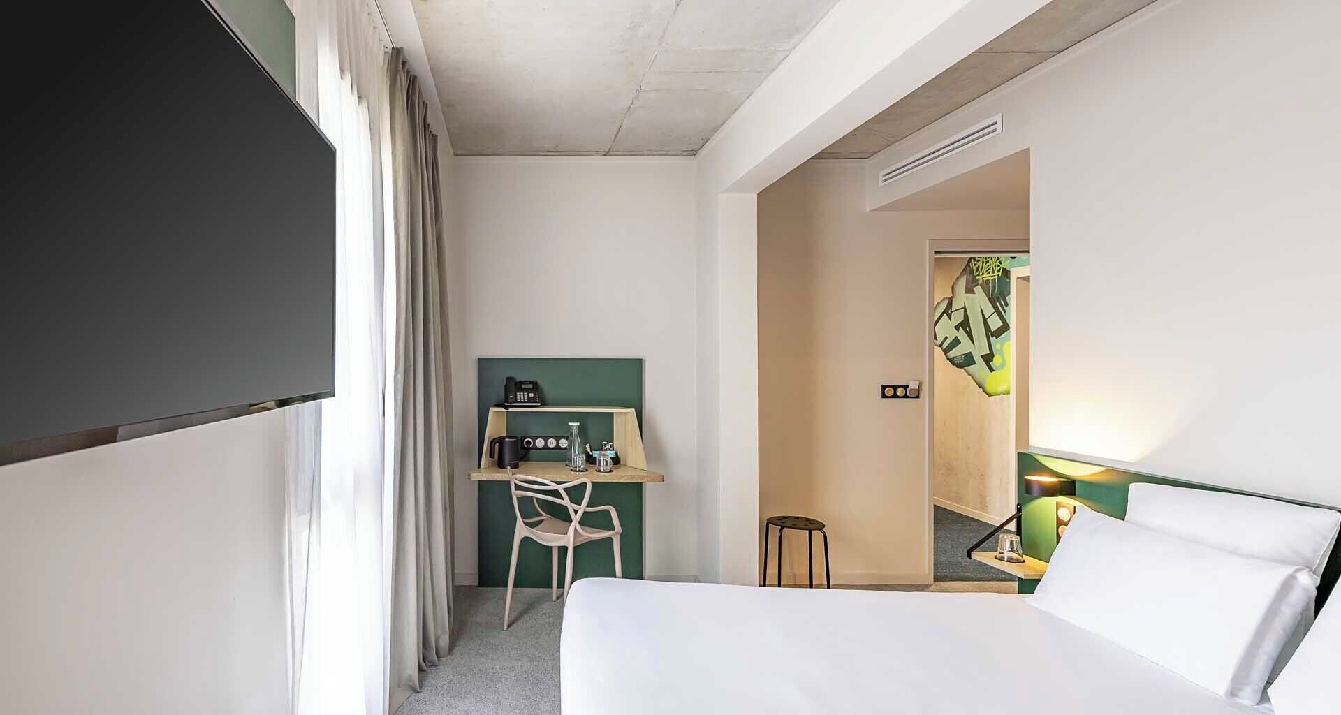 A hotel room with a bed, a desk with a telephone and coffee maker, and a small stool.