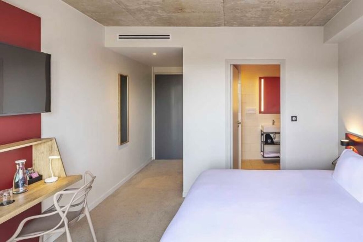 A hotel room with a bed, a desk with a chair, a TV mounted on the wall, and a bathroom visible through an open door.