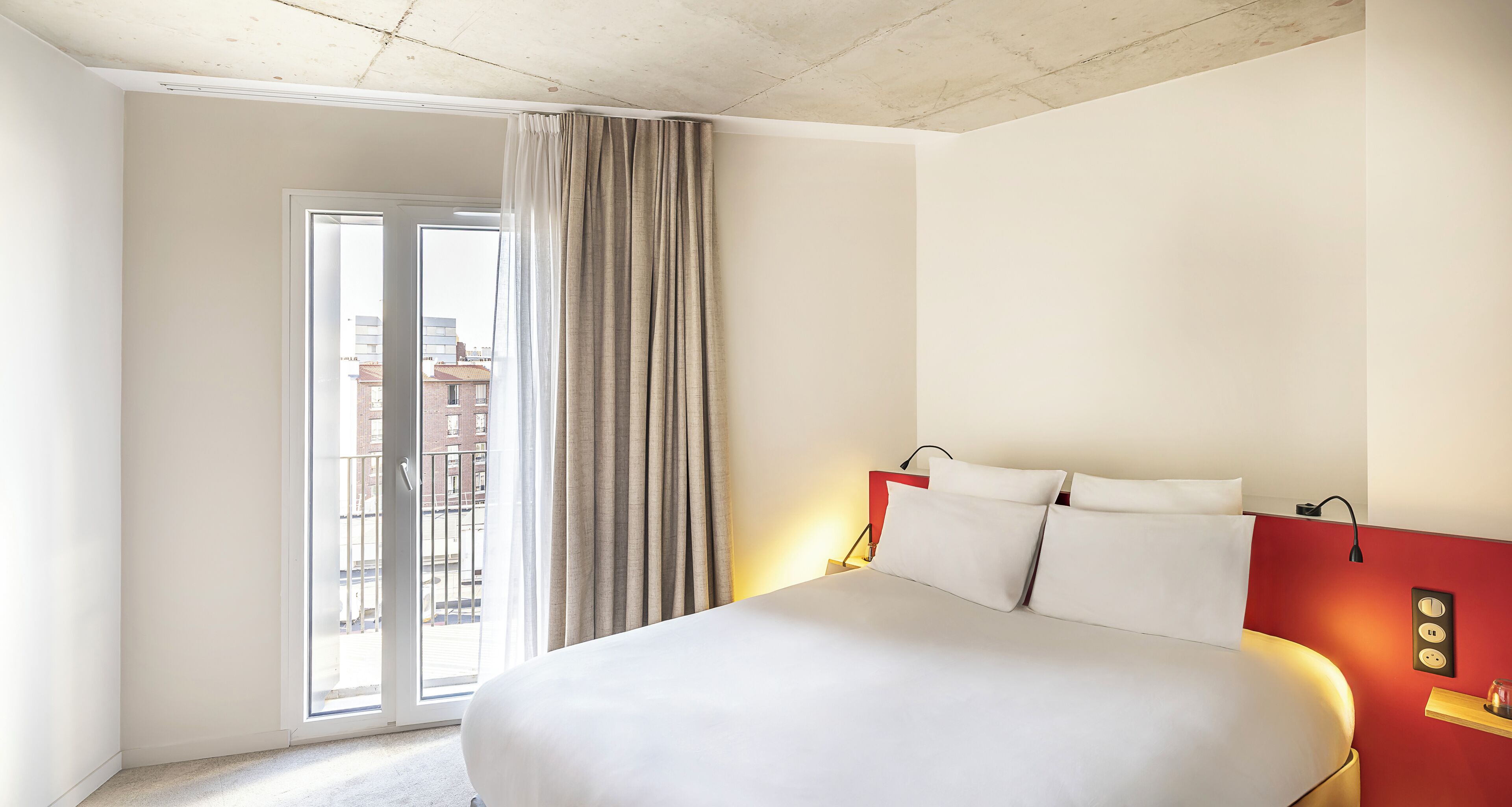 Une chambre d’hôtel avec un grand lit, une tête de lit rouge, une porte-fenêtre coulissante et une vue sur les bâtiments environnants.