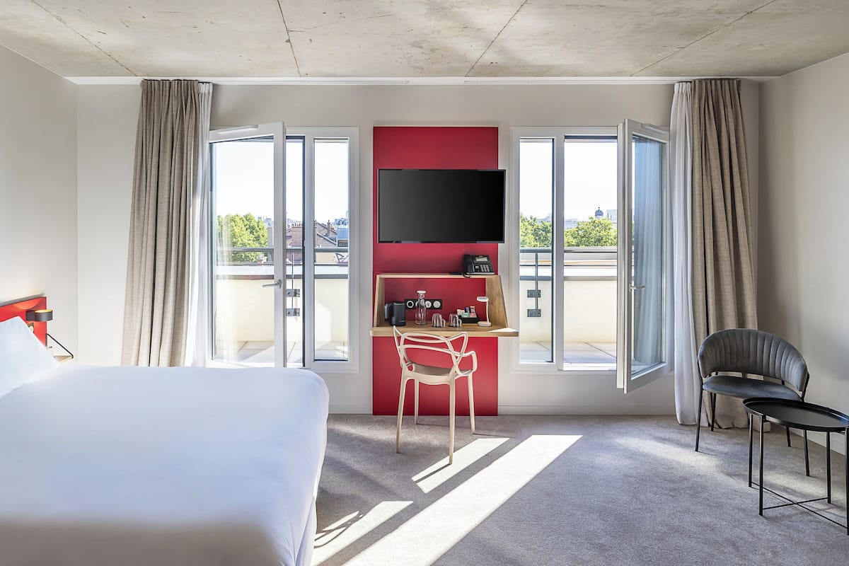 A hotel room with a bed, a desk with a chair, a TV, and large windows.