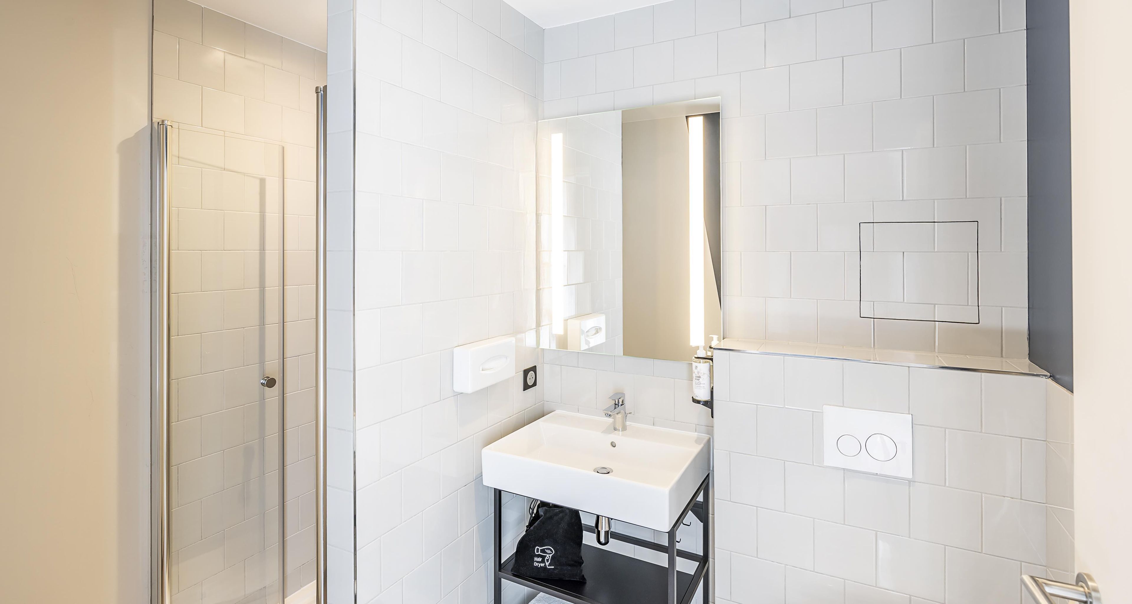 Ein modernes Badezimmer mit weißen Fliesen, einer Glasdusche, einem weißen Waschbecken und einem Spiegel mit integrierter Beleuchtung.