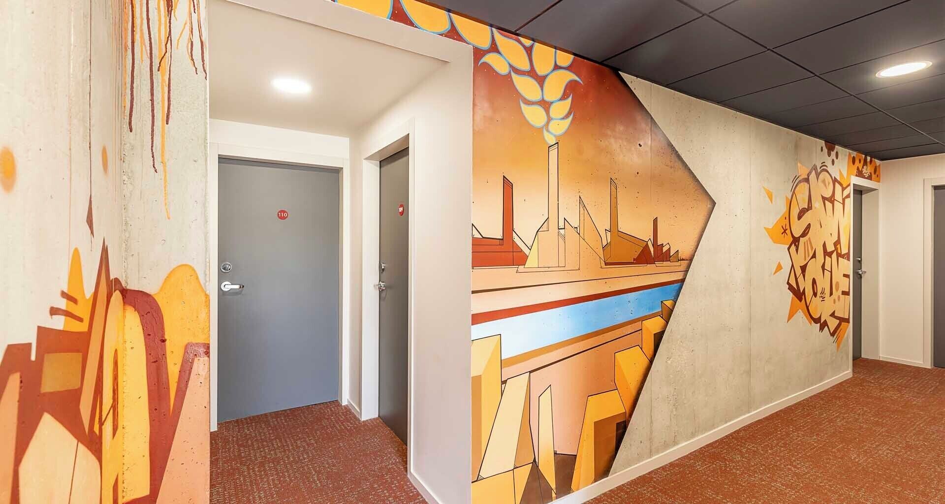 Un pasillo con un mural que representa una escena industrial, una puerta con el número 110 y suelo cubierto de alfombra.