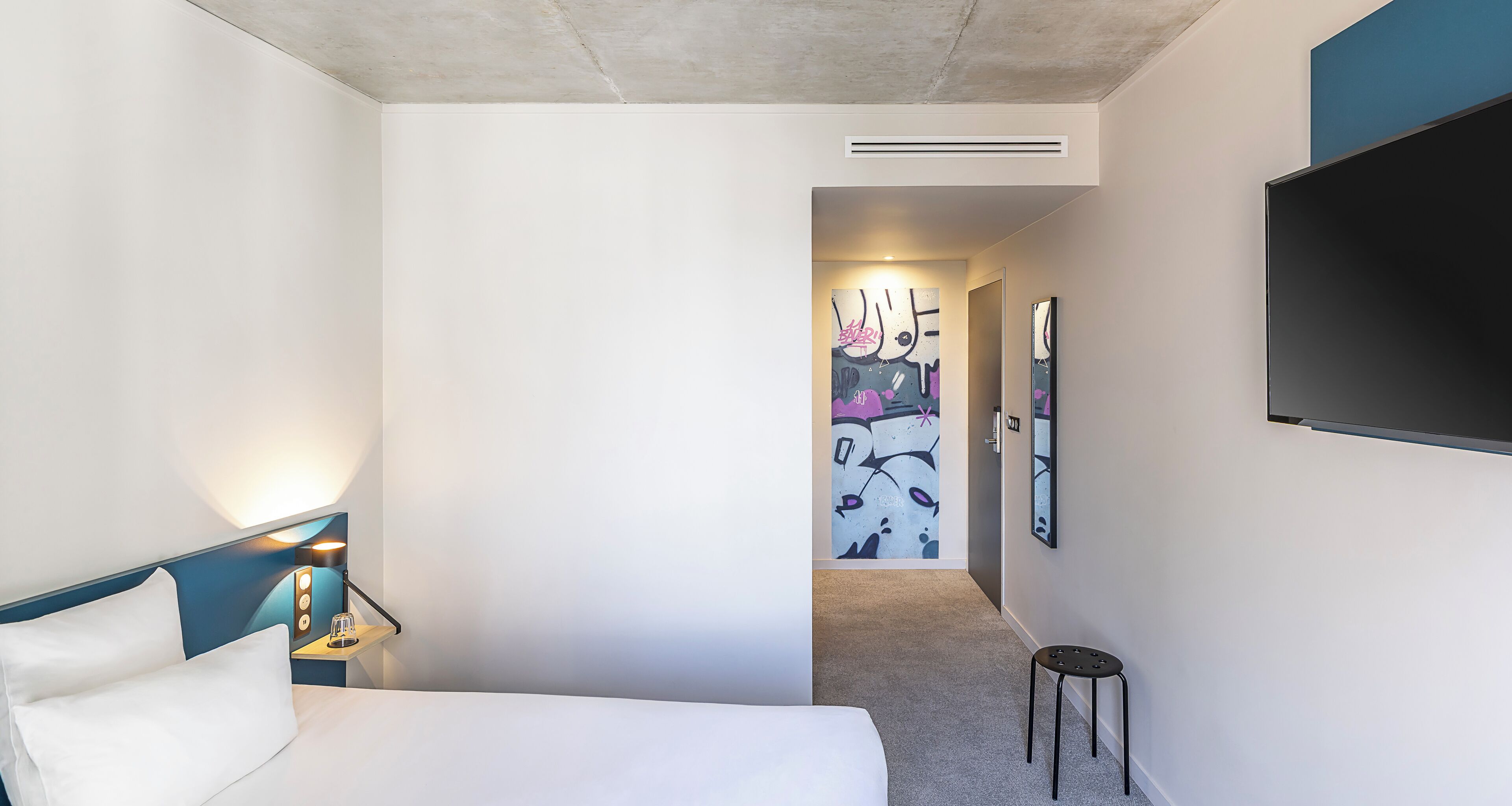 A hotel room with a bed, bedside lamp, wall-mounted TV, and a small stool.