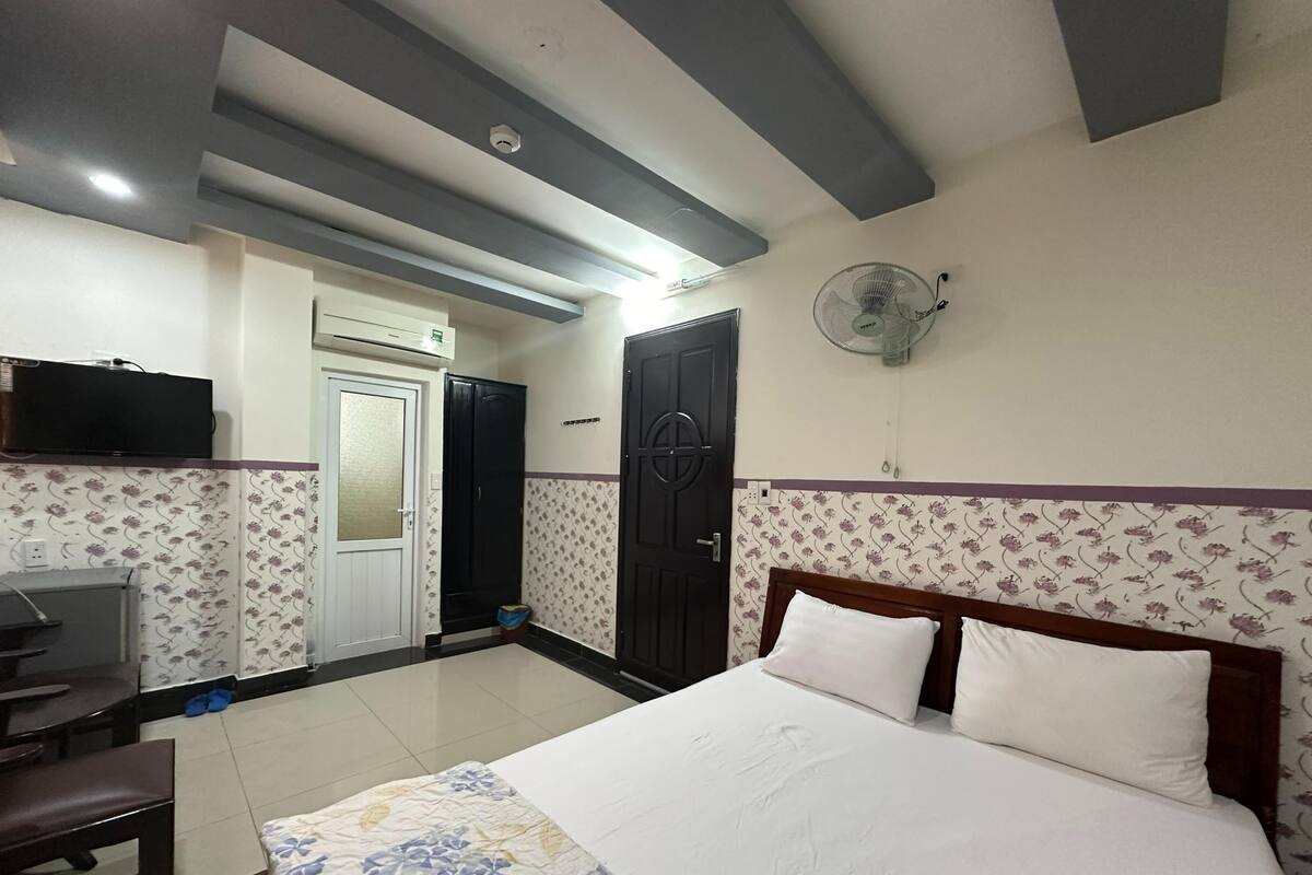 A hotel room with a bed, a desk, a chair, a TV, and a wall-mounted fan.