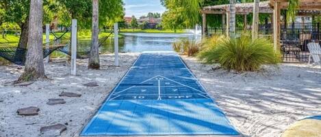 A shuffleboard table with a blue surface and numbered scoring areas, surrounded by trees and a body of water.