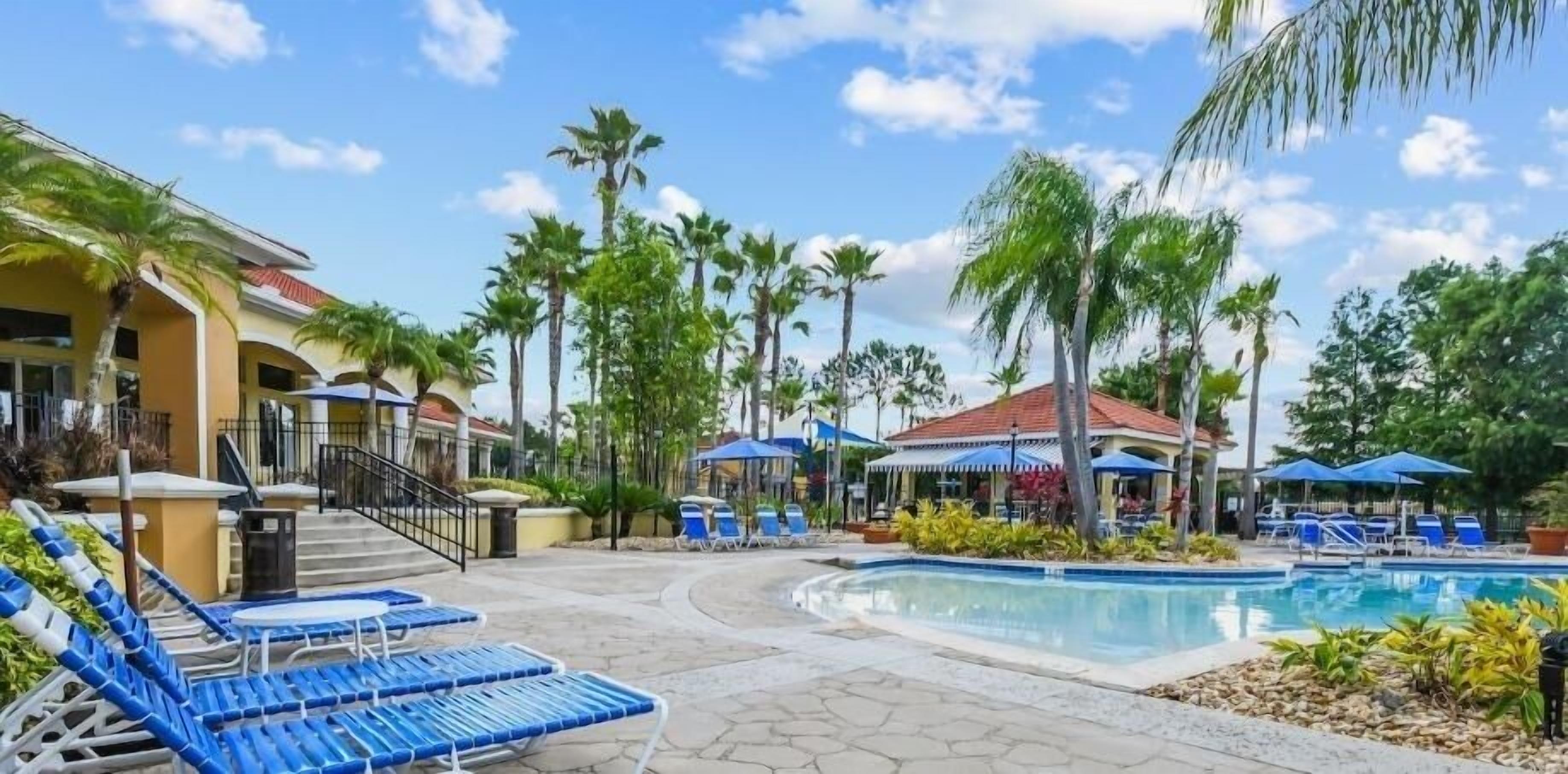 A poolside area with lounge chairs, a swimming pool, palm trees, and a building with a red roof.