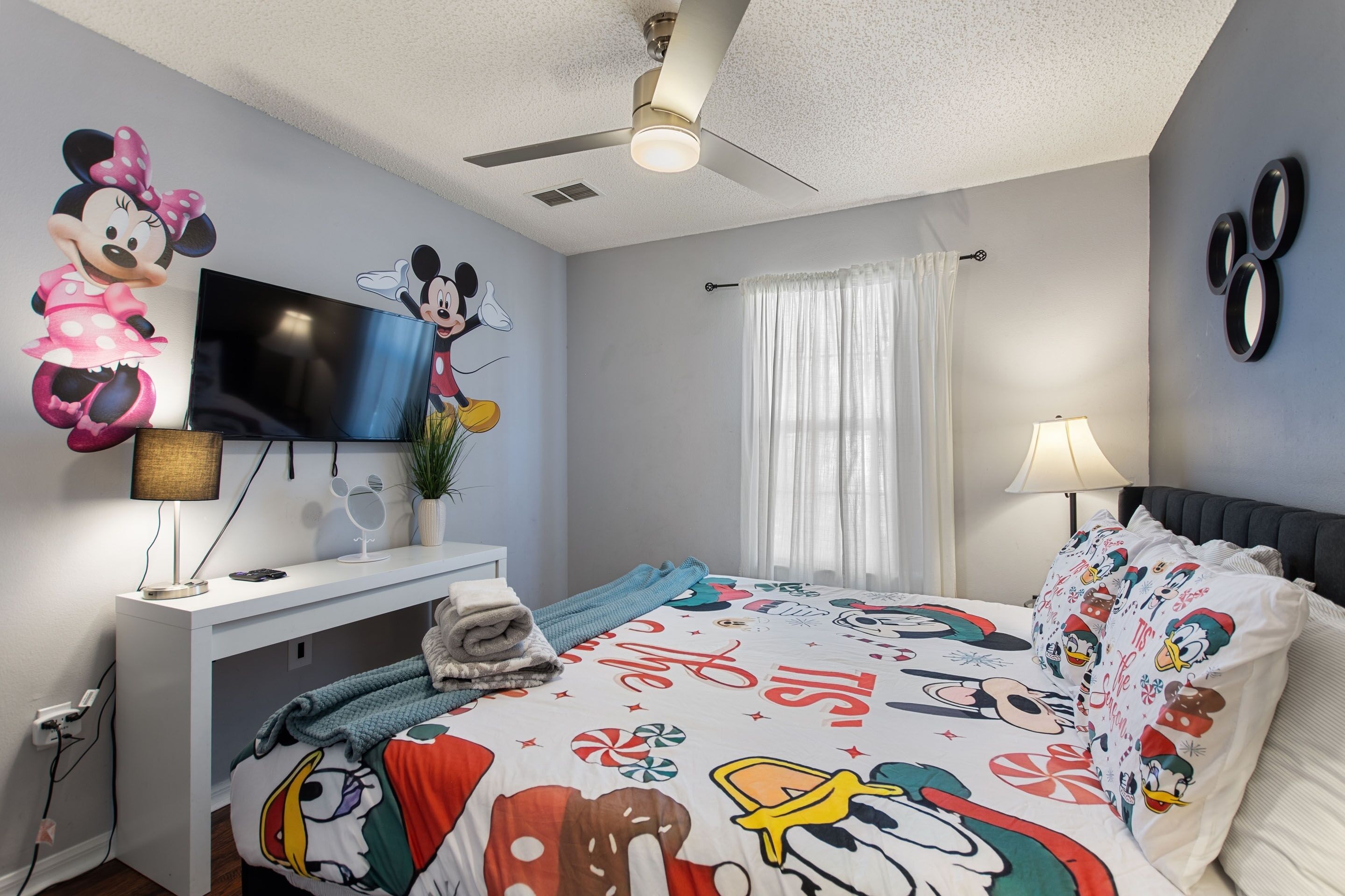 A bedroom with a bed featuring a Mickey Mouse themed bedspread, a flat-screen TV on a white stand, a ceiling fan, and a window with white curtains.