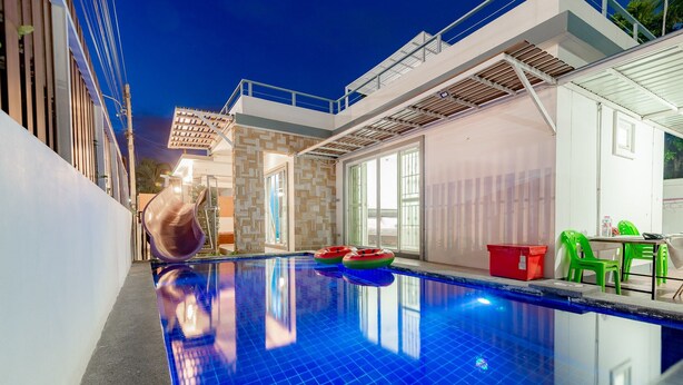 A swimming pool with a slide, a red trash bin, and green chairs.