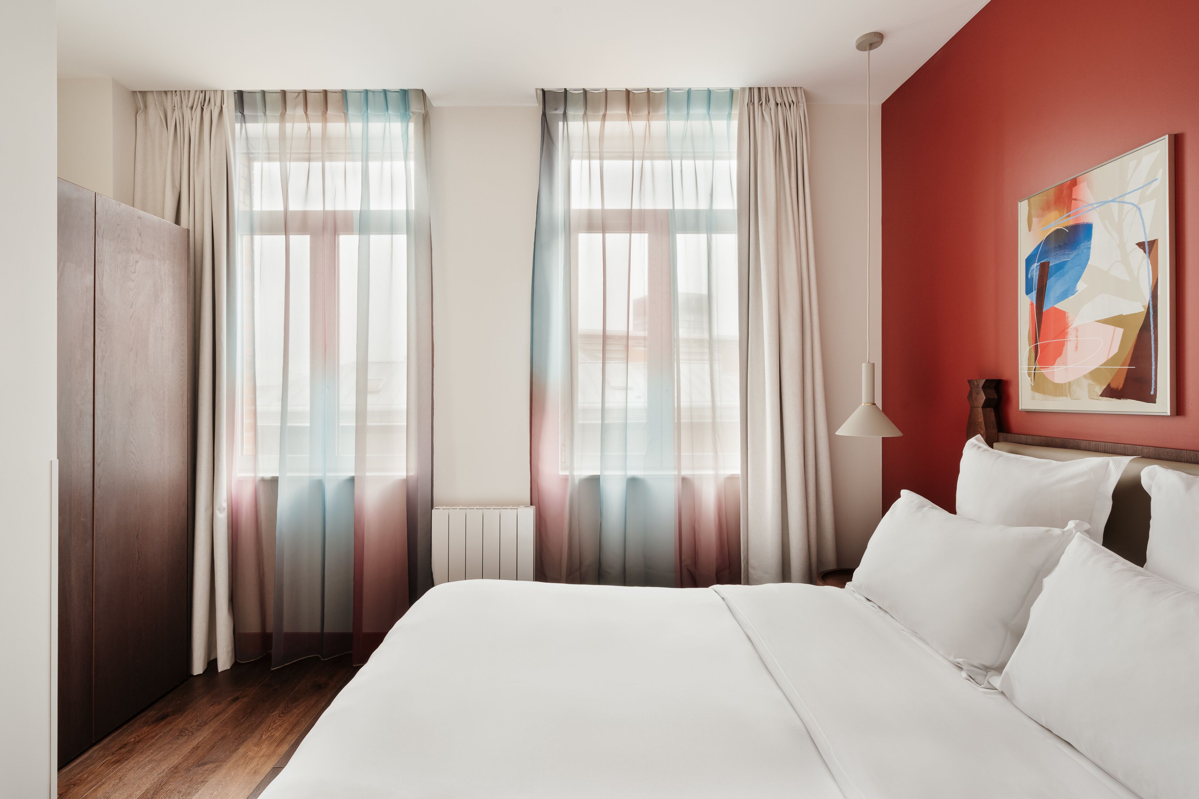 Een hotelkamer met een bed, gordijnen, een radiator, een lamp en een schilderij aan de muur.