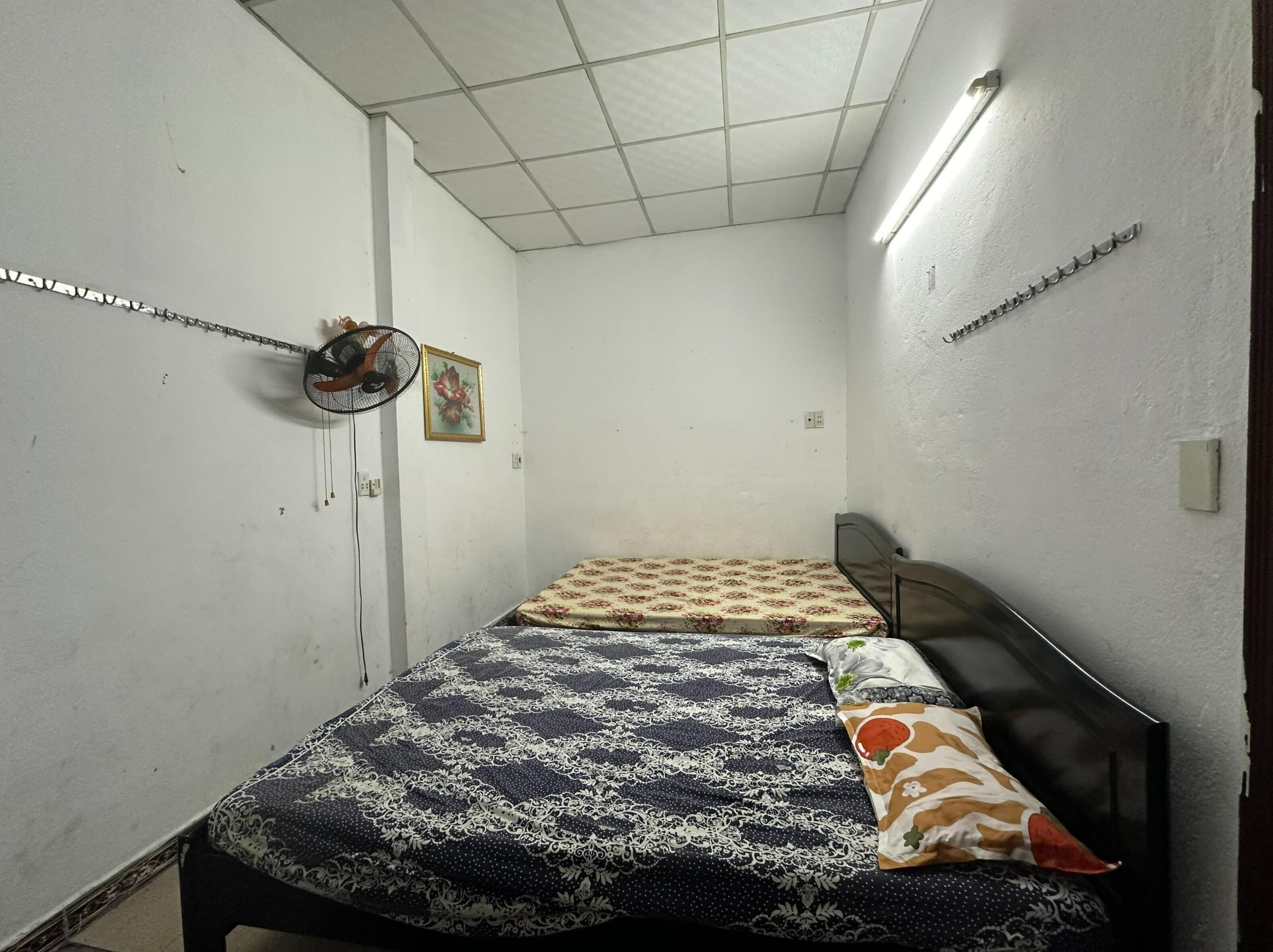 A small, dimly lit room with two single beds, a fan, and a small picture on the wall.