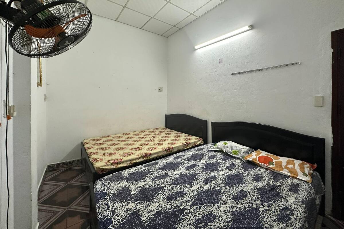 A small, sparsely furnished room with two beds, a ceiling fan, and a fluorescent light.